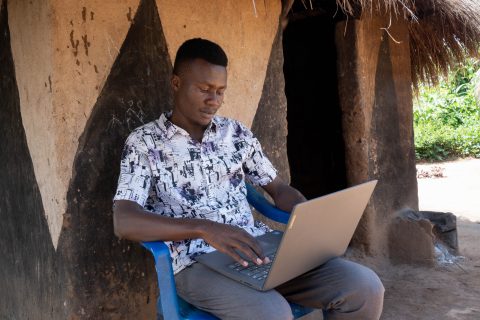 Richard Luate sitting on a plastic chair outside his hut in the Bidi Bidi refugee camp, with a computer in his lap.