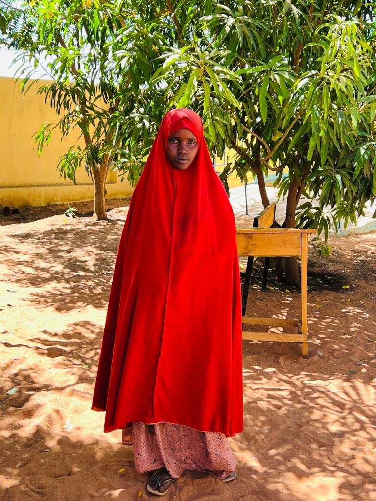A girl in a red Abaya stands in the indoors in front of a schooldesk. 
