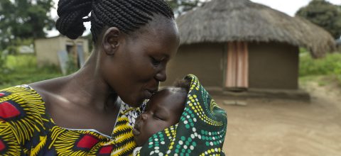 Nainen suutelee vauvan otsaa Etelä-Sudanissa. Taustalla näkyy savesta ja ruohosta tehty rakennus.