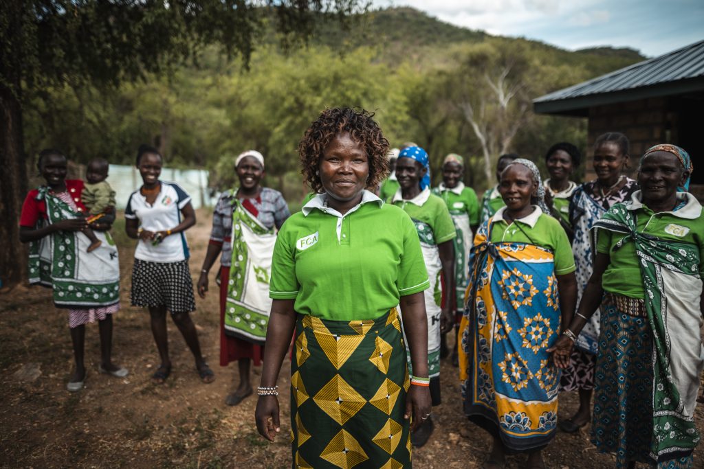 A woman in a green shirt with the FCA logo stands in front of a number of other women in similar shirts. They are outside and wearing traditional east African wraps. 