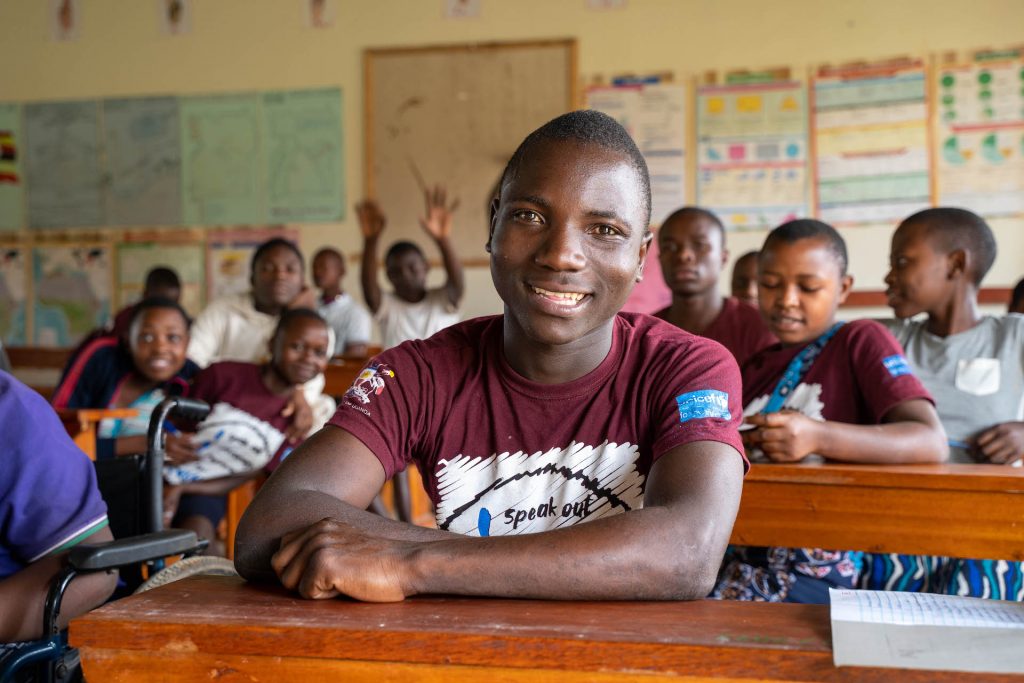 A Ugandan boy in sitting at a desk in a classroom, with classmates behind him.