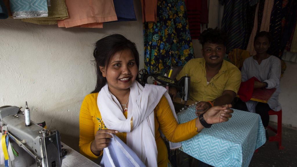 A woman and a man sit at tables with sewing machines posing for the camera