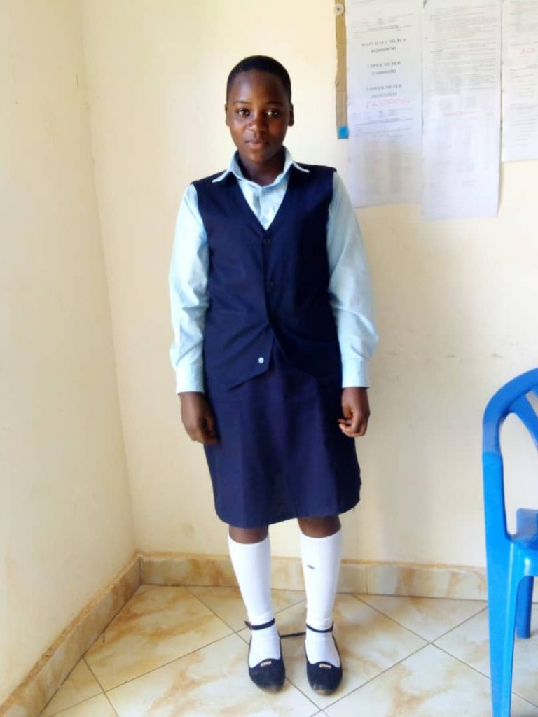 A girl in school uniform stands in the corner of a classroom looking at the camera.
