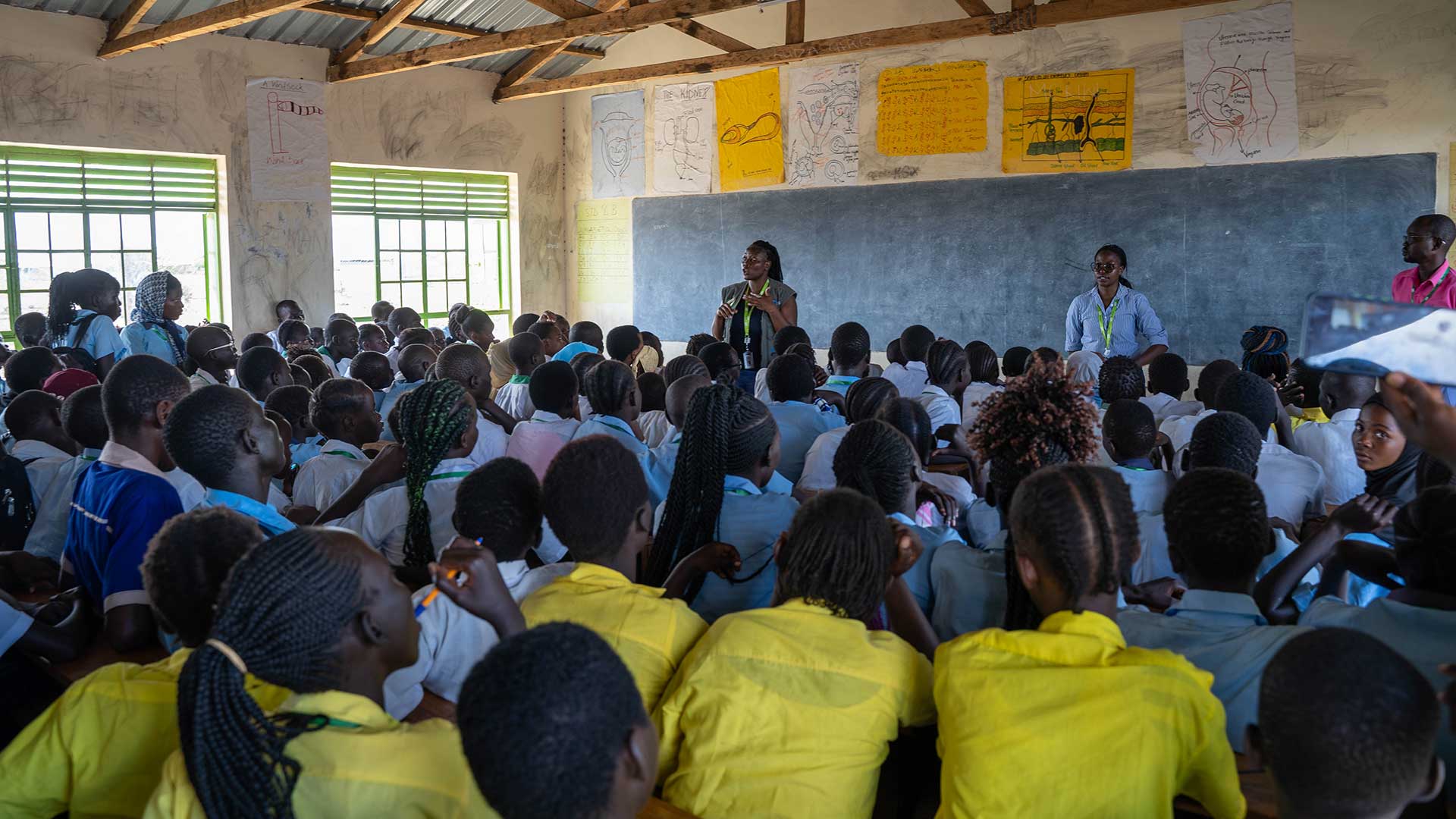 A full class of students sit in a class room in Kenya. There is a teacher in front of the class.