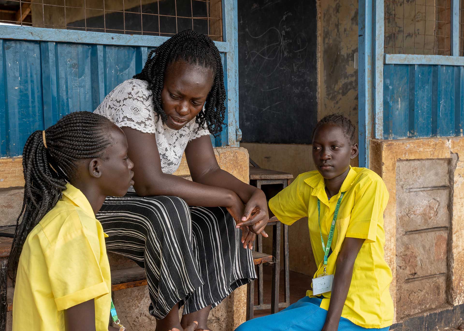 A woman and two school girls sit on a porch in front of a building in Kenya.