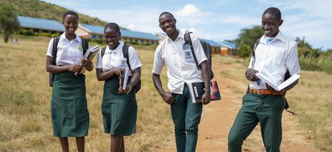Four teenagers in school uniform stand outside holding their books and smiling at the camera