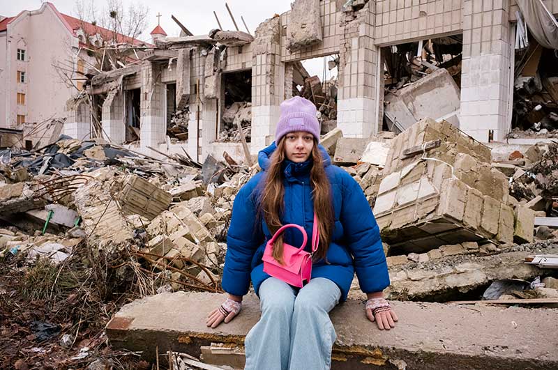 A teenage girl sits in front of a damaged building in Ukraine.