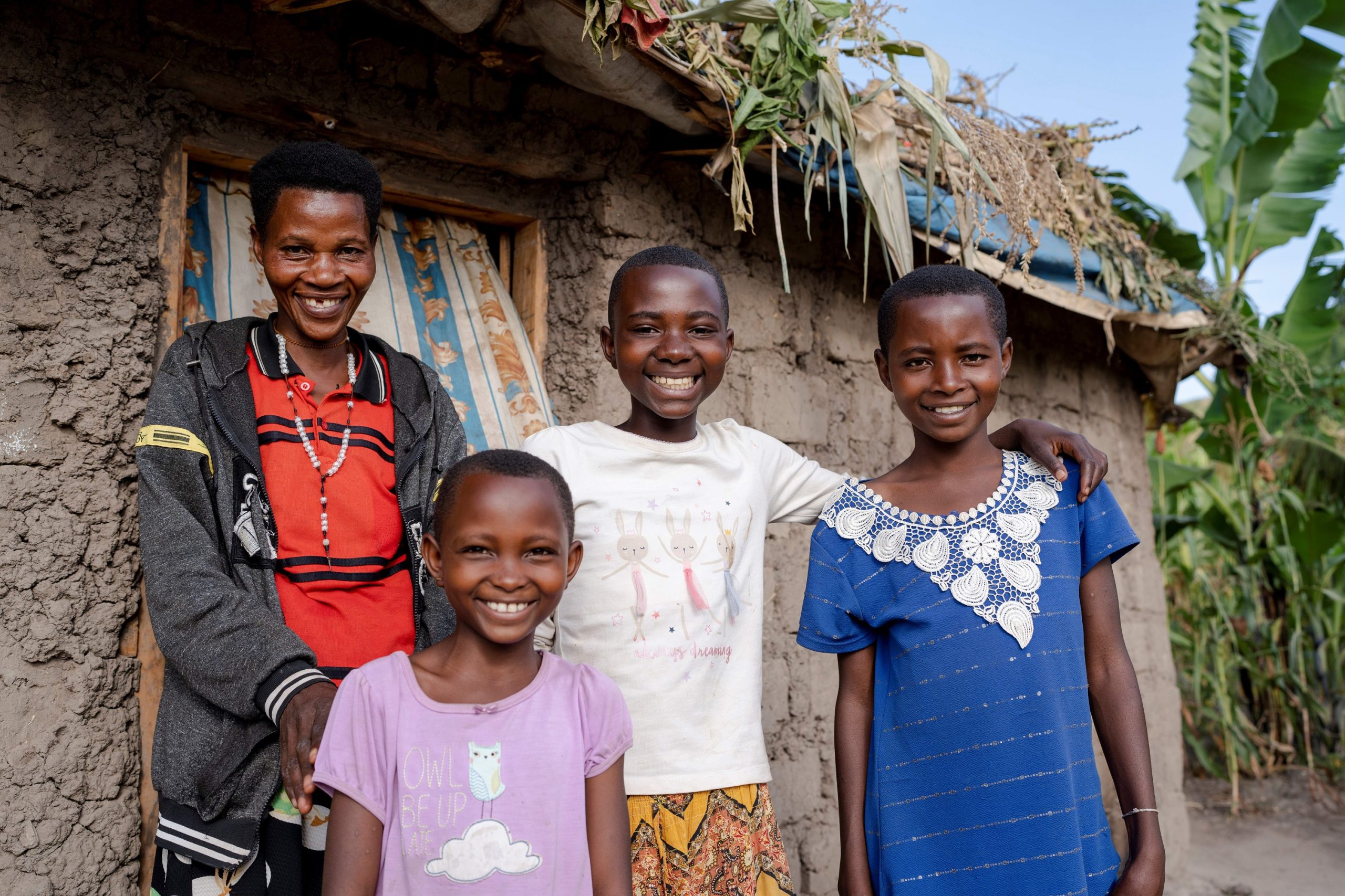 A woman and three girls stand in front of a small hut smiling at the camera