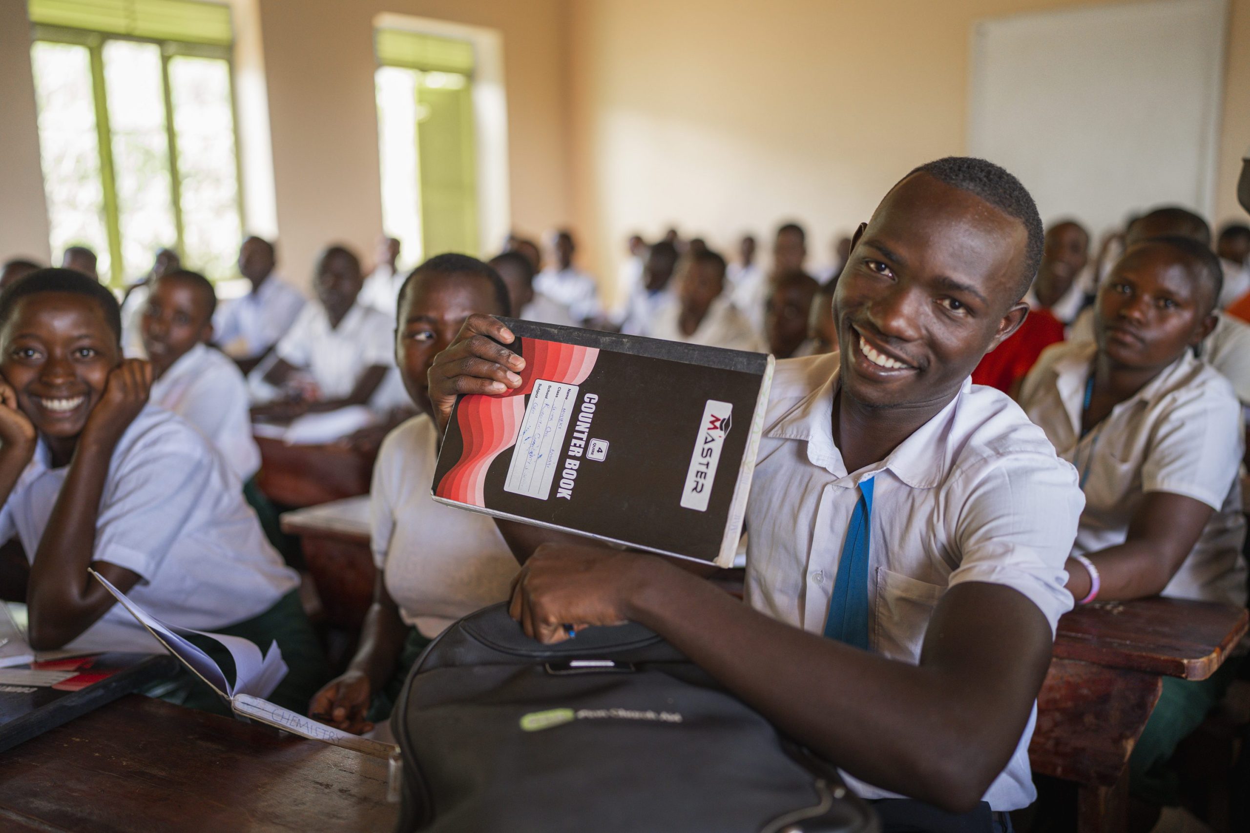 A teenaged boy in school uniform sits at a desk in a full classroom and holds up a text book to the camera. He is smiling.