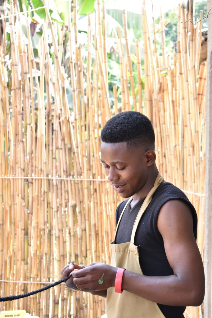 A man stands in front of a straw fence and braids a long pony tail. He is wearing an apron and has a look of concentration