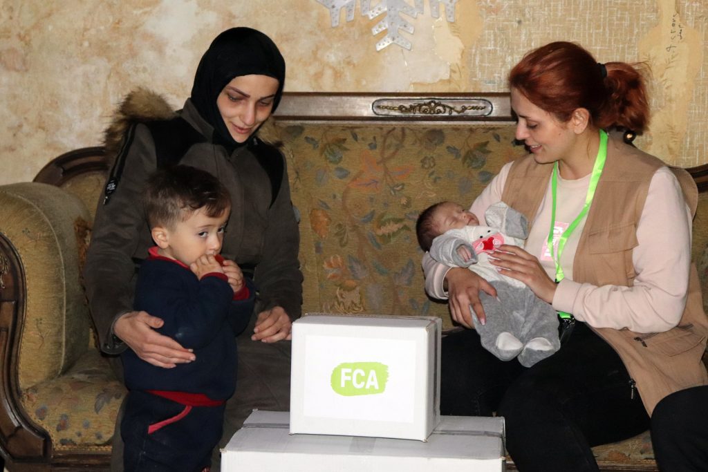 Two women sit on a coach. One has her arm around a small child while the other is holding a newborn baby. A box with an FCA logo sits in front of them.