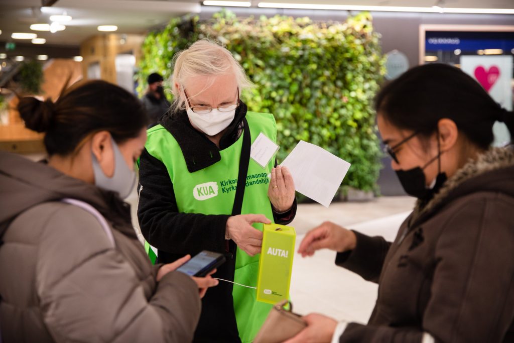 A woman in a mask and an FCA branded vest accepts cash donations from two other women in the street.