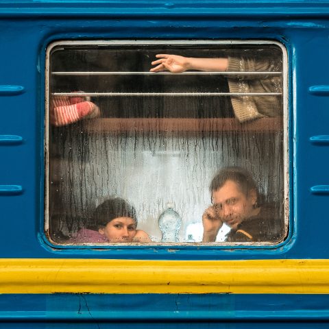 A couple look out at us through a steamed-up train window. In the luggage rack above them lies a child. The train's colours are the blue and yellow of the Ukrainian flag