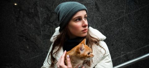 A woman in a beanie hat and coat holds a ginger cat closely to her chest. She is looking pensively off camera.