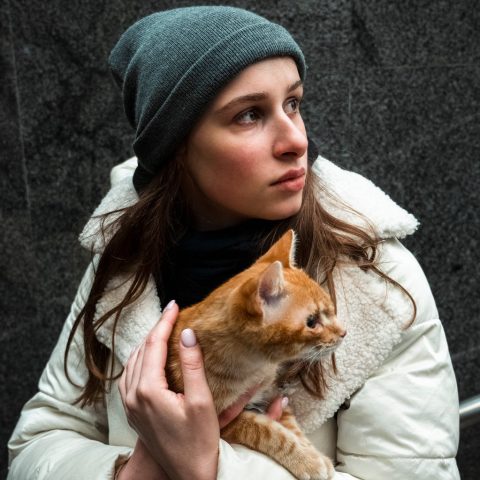 A woman in a beanie hat and coat holds a ginger cat closely to her chest. She is looking pensively off camera. 