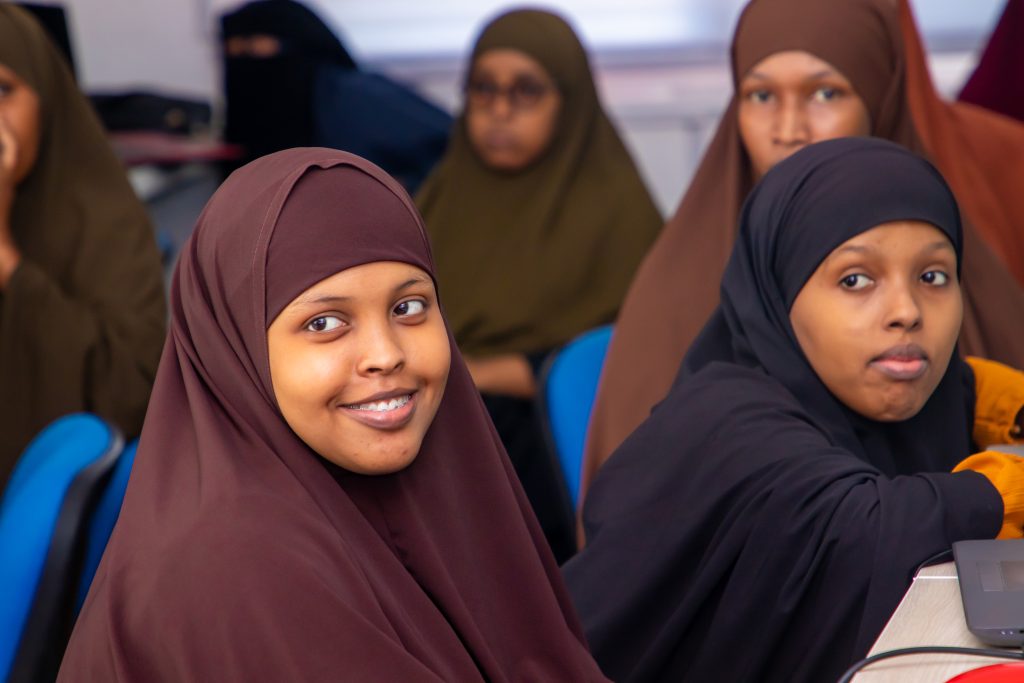 Two young women in hijabs and abayas sit next to computers. They are looking towards the camera and smiling