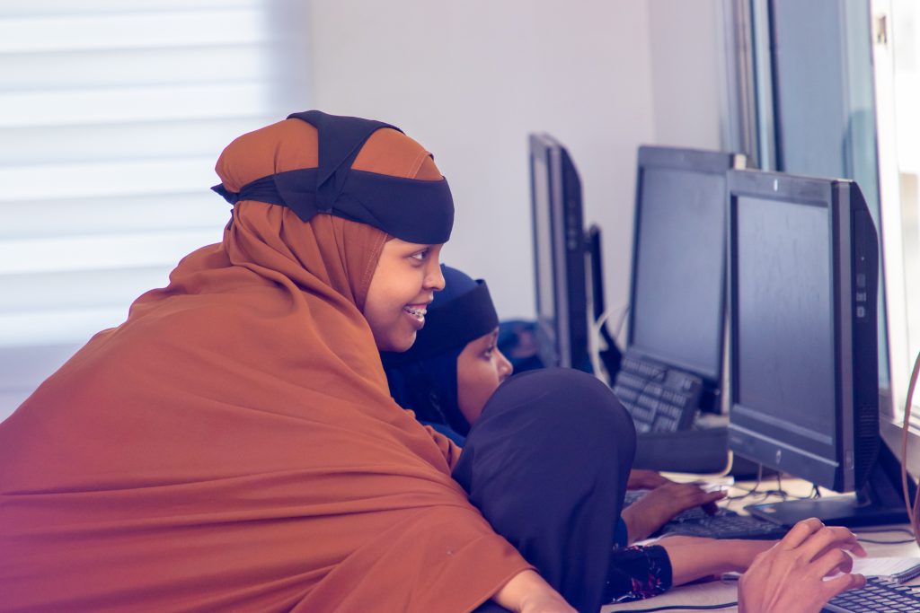A woman in an abaya an hijab leans over a computer. She is smiling. 