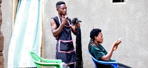 A man stands holding a long braid of hair, while a woman sits on a chair in front of him looking at a mobile phone.