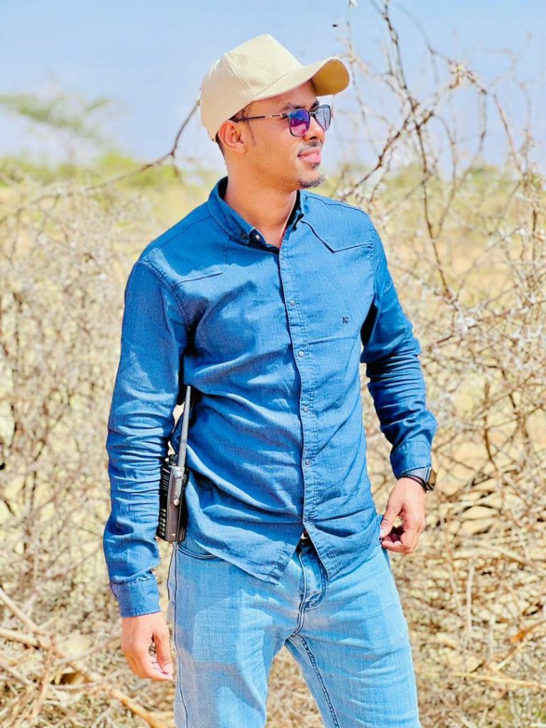 A smiling man in a baseball cap, sunglasses and denim shirt and jeans stands in an arid landscape