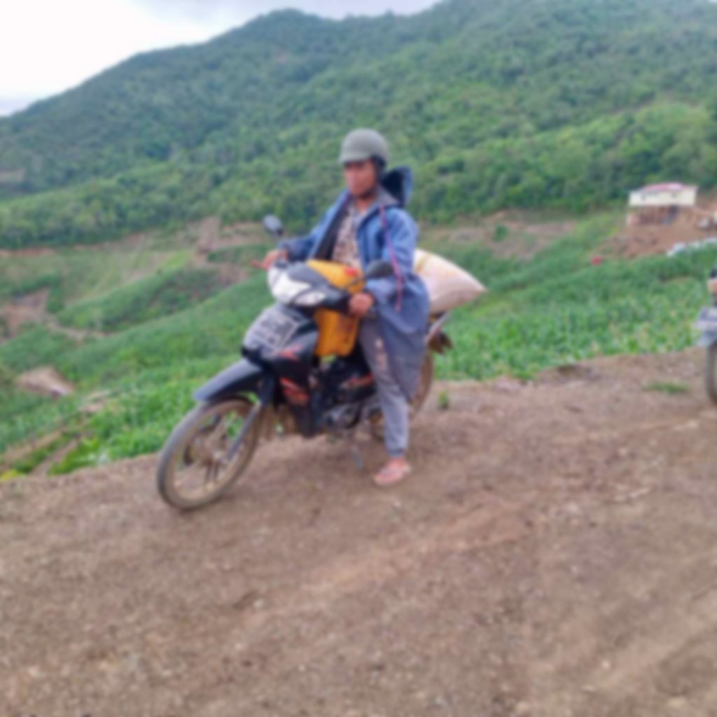 A man sits on a motorbike atop a ridge overlooking a shallow valley. His features are blurred.