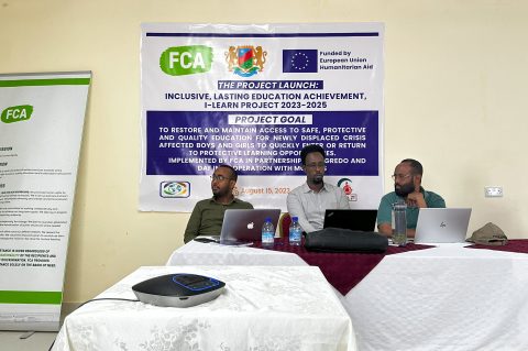 Three men sit at a desk in an office in front of a large poster with details about the FCA iLearn project