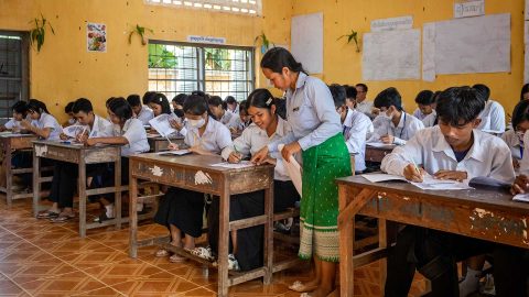 Oppilaat istuvat pulpeteissaan kambodzalaisessa koulussa. Opettaja auttaa eturivissä istuvaa tyttöä.