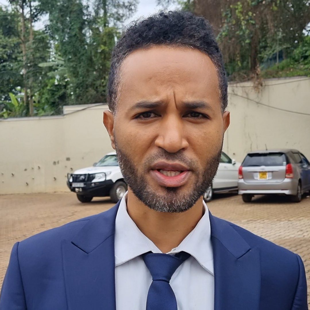 A young man in a suit and tie stands in a car park looking into the camera with a serious expression