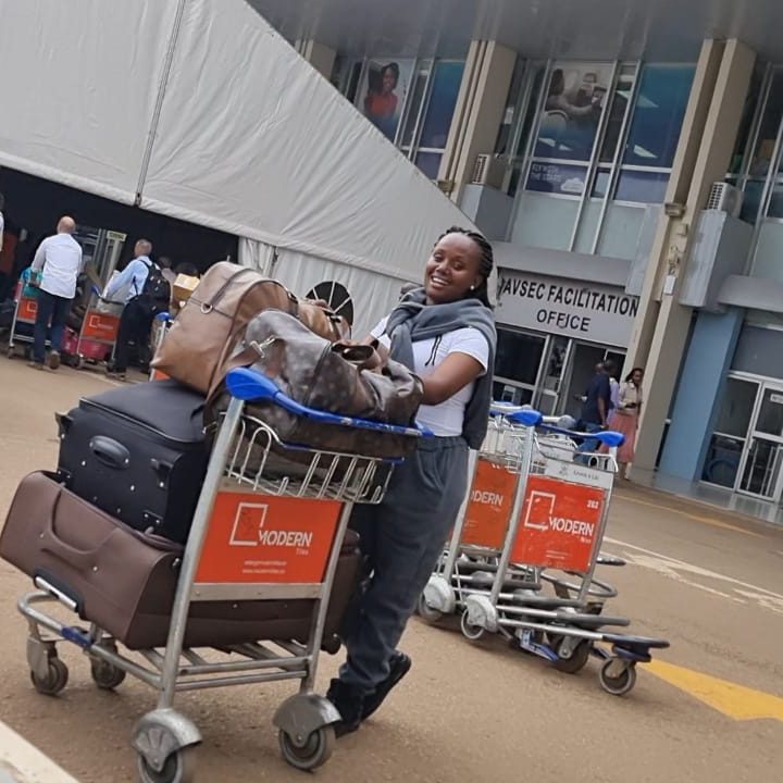 A smiling woman stands with a trolley full of luggage in front of an airport building. She is smiling.