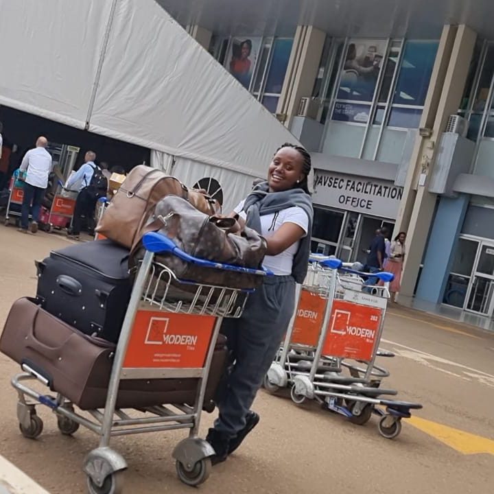 A smiling woman stands with a trolley full of luggage in front of an airport building. She is smiling.