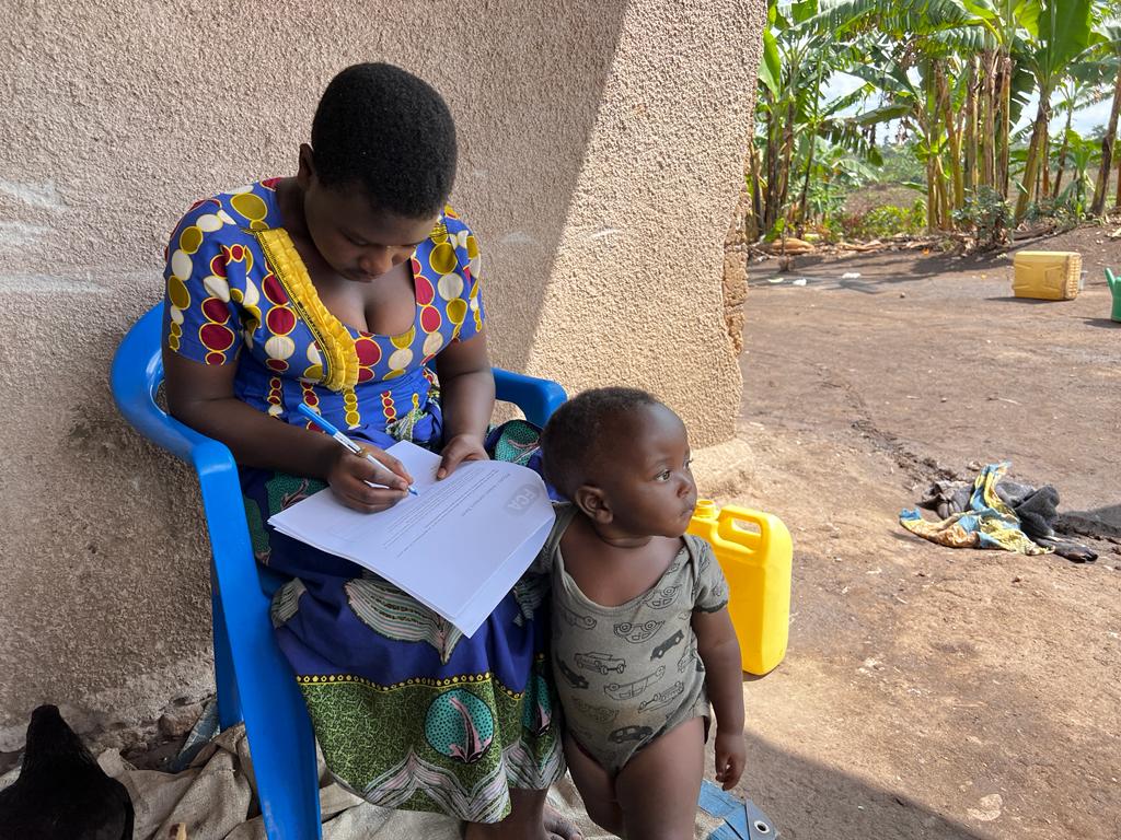 A girl in a brightly printed dress sits outside on an plastic chair writing into a book, while a small child stands by her legs
