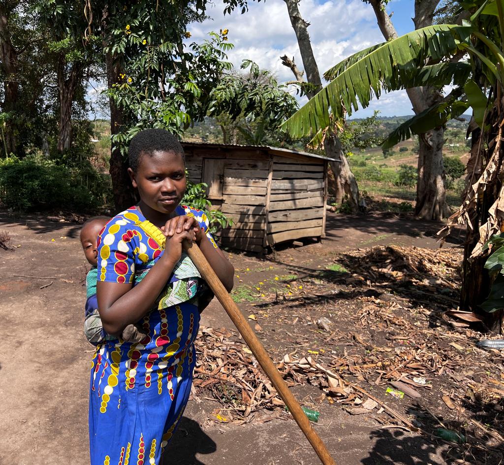 A girl in Ugandan national dress stands in her garden, holding a garden tool in her hands. She has a baby in a sling on her back.