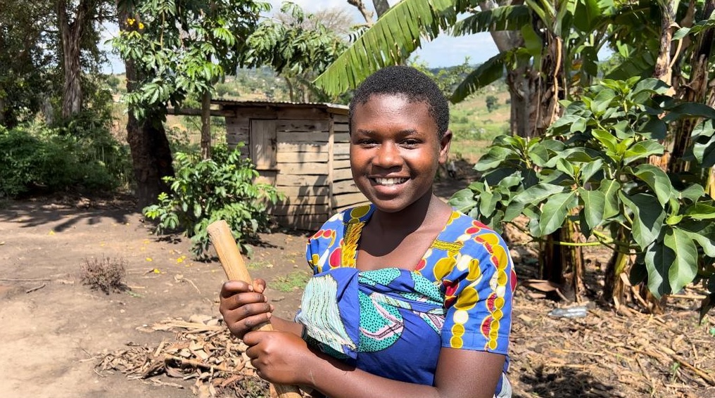 A smiling girl in Ugandan national dress stands in her garden, holding a garden tool in her hands