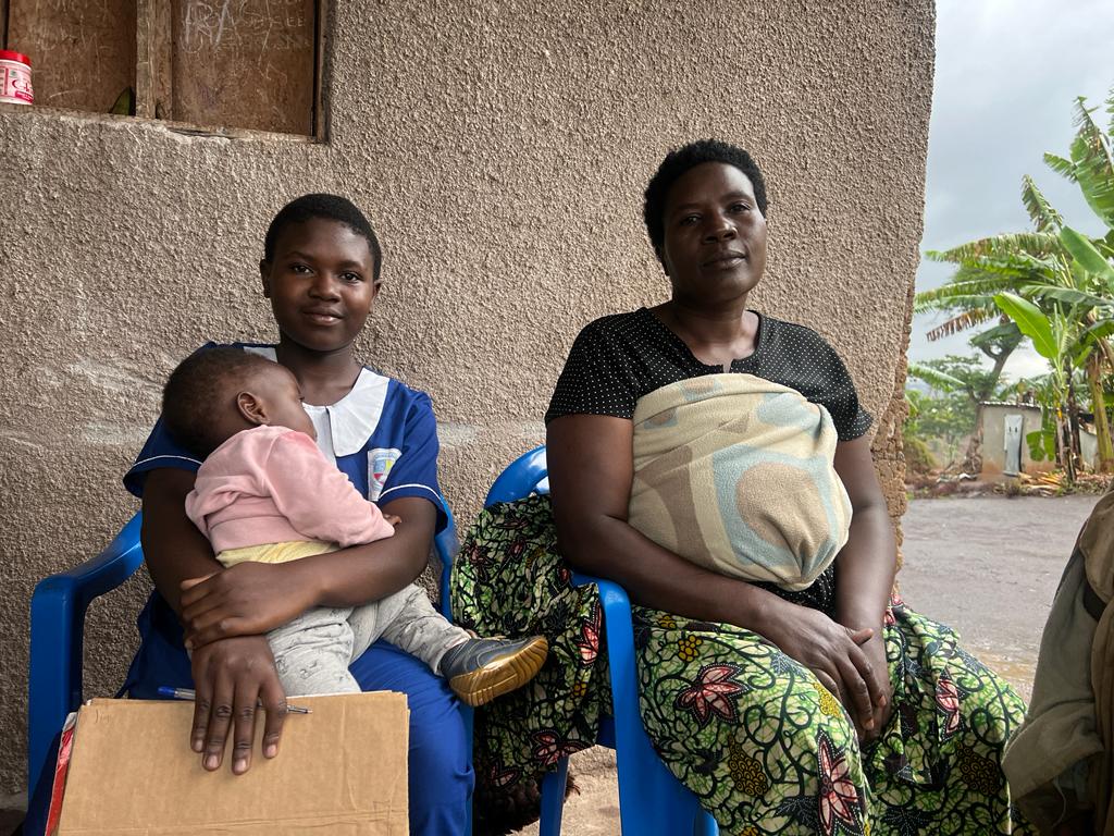 A girl with a small child on her lap sits in a plastic chair next to an older woman. They are outside a house. Both are softly smiling