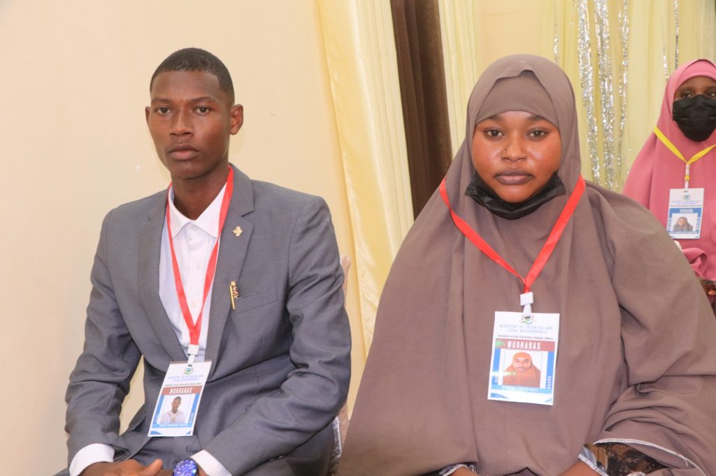 A man and a woman sit together in a room with IDs around their necks. Both wear serious expressions