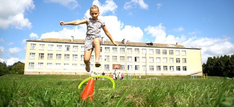 A girl jumps over a hoop in a field of grass outside a large school building