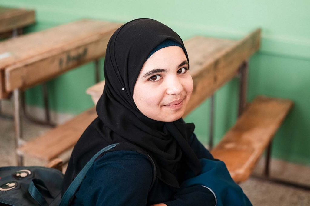 En syrisk tjej tittar på kameran och ler i ett skolklassrum.