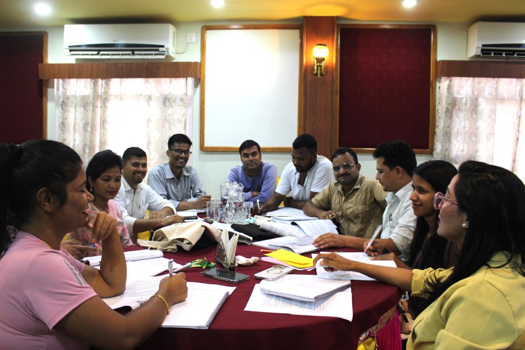 A group of people sit around a circular table in a small office, talking and laughing.