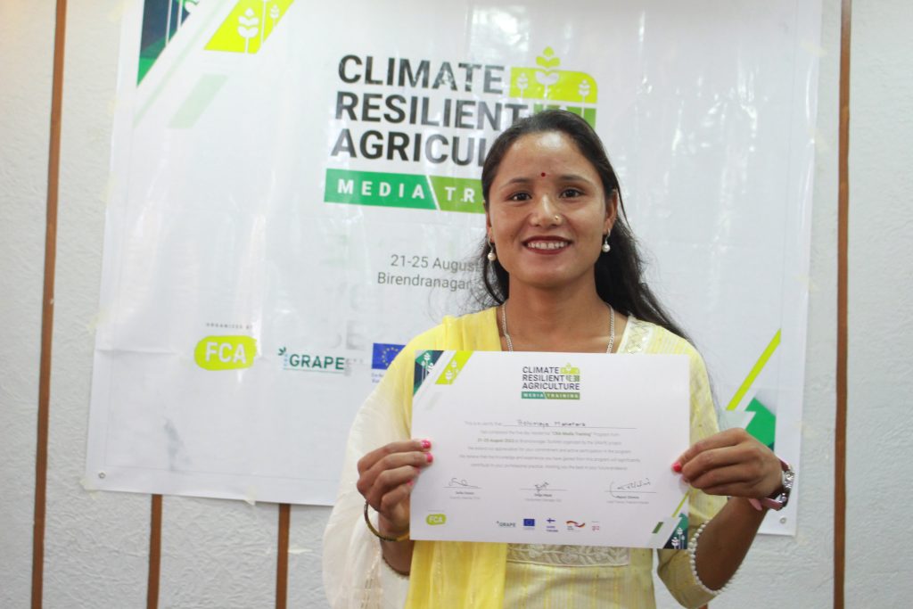 A smiling woman holds up a certificate, while standing in front of a banner that reads "Climate Resilient Agriculture media training".