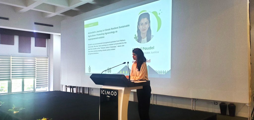 A woman stands at a lectern and talks into a microphone. Behind her a presentation shows her career background in Climate Resilient Agriculture.