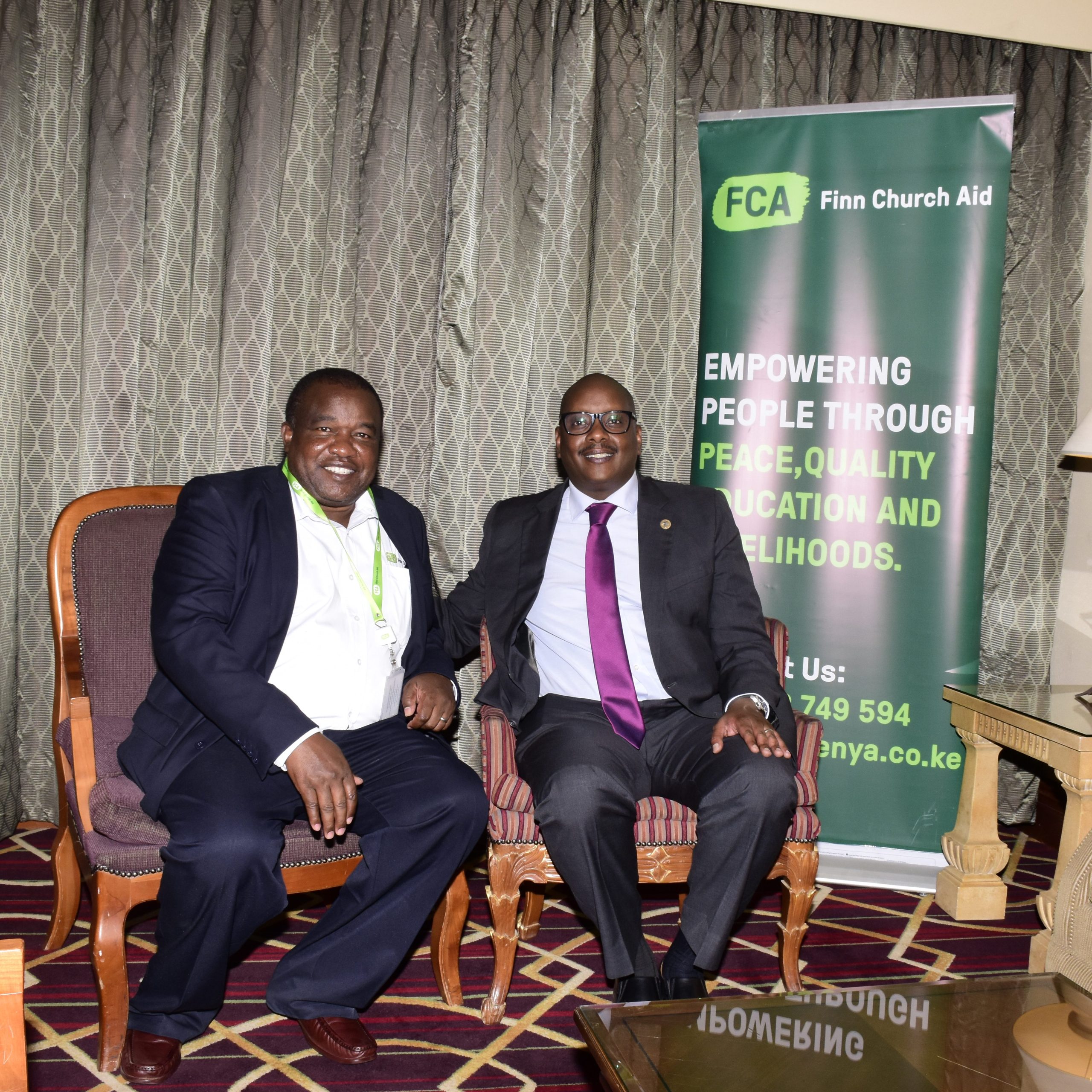 Two men sit in an office and smile at the camera. Behind them is a banner bearing the FCA logo