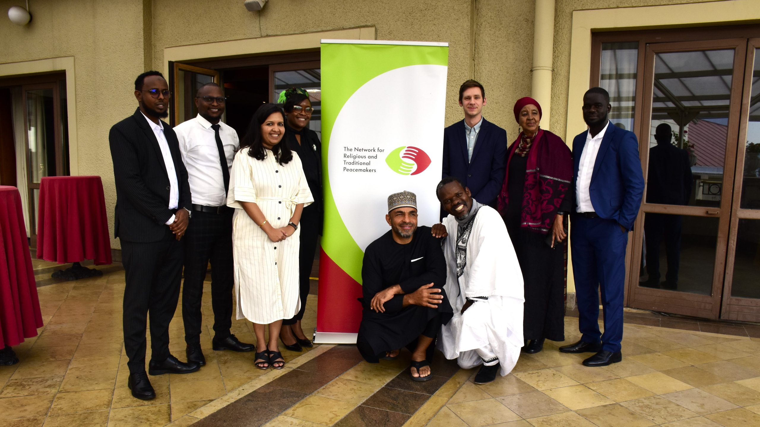 A group of people pose for a photo outside. Behind them is a banner that bears the logo of The Network for Religious and Traditional Peacemakers