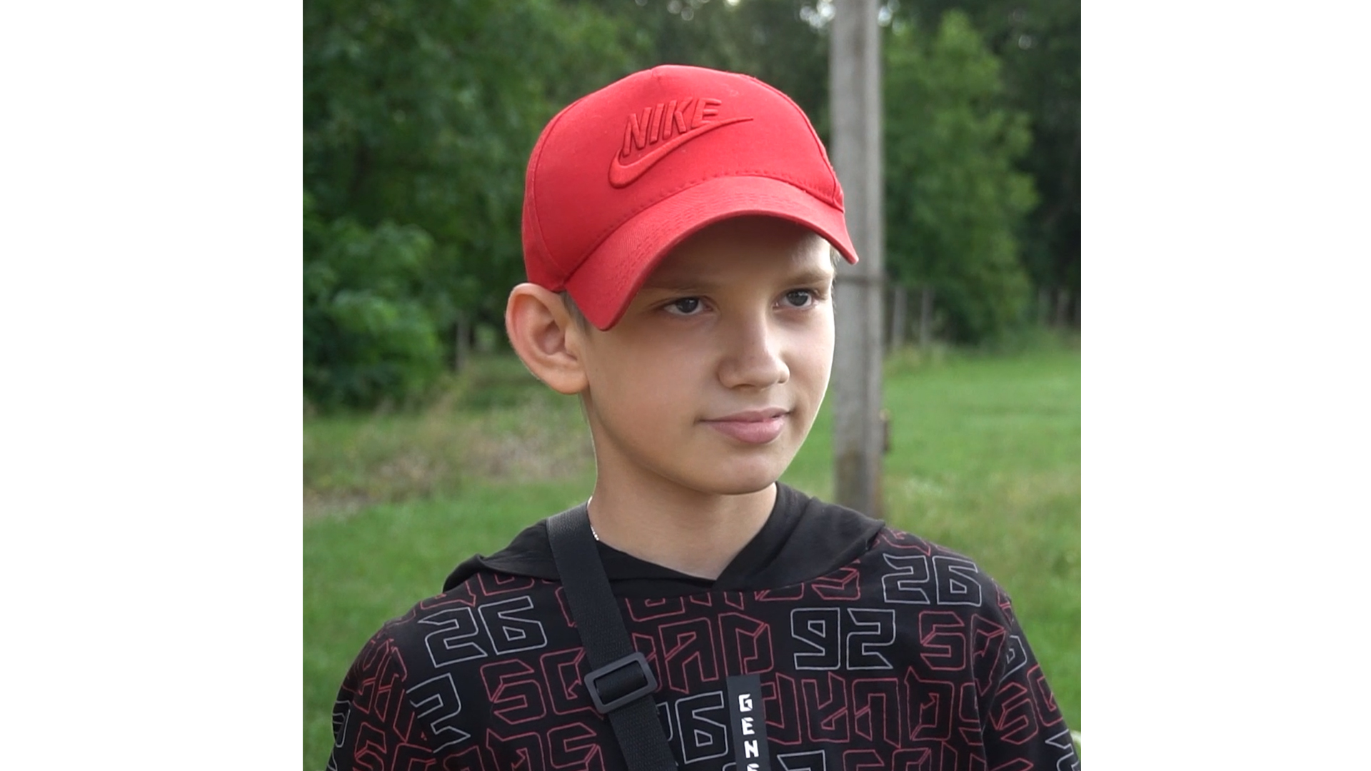 A boy in a patterned sweater and a red baseball cap stands outside in a field and looks just past the camera with a calm expression