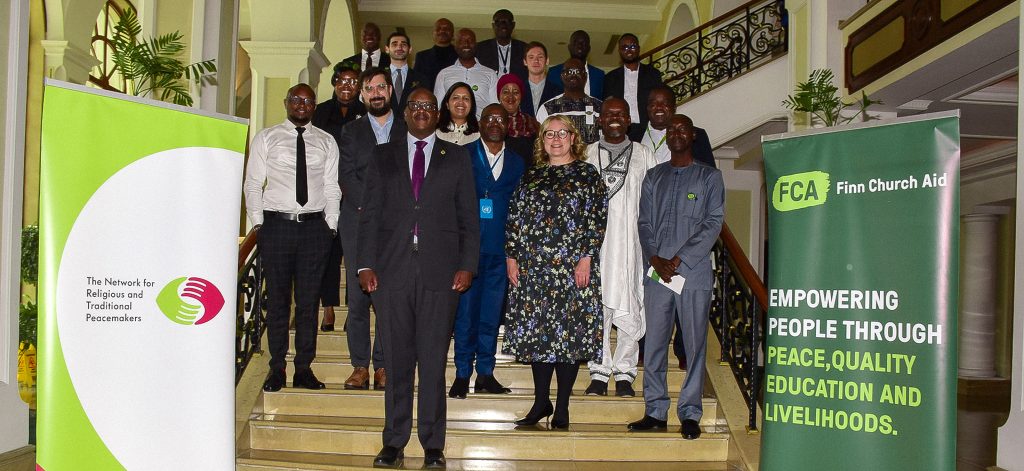 A group of people pose for a photo on an indoor staircase. Either side of them are banners showing logos of FCA and The Peacemakers Network