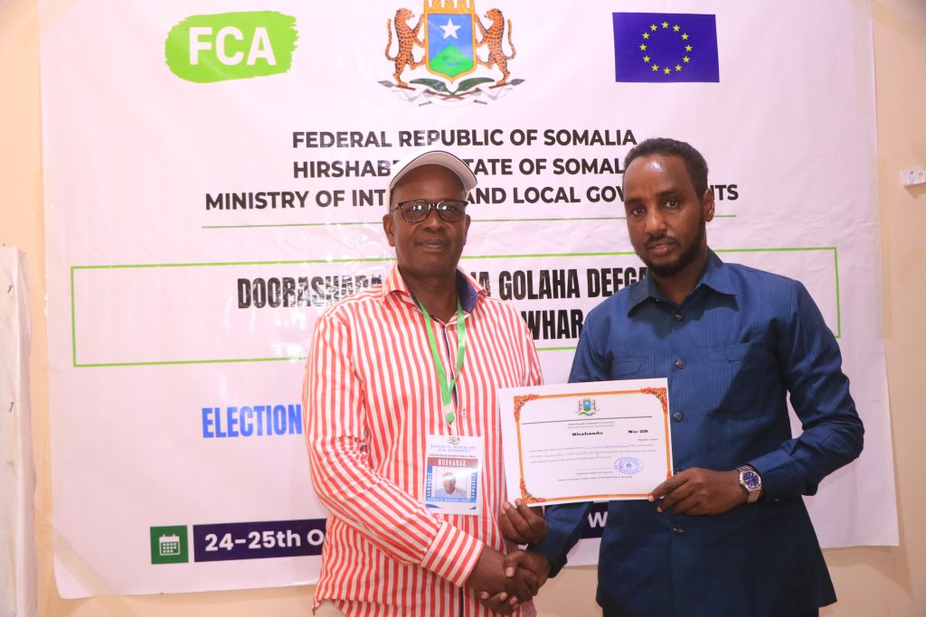 Two men shake hands standing in front of a banner bearing the logos of FCA, the Federal Republic of Somalia and the European Union. One of the men is holding a certificate