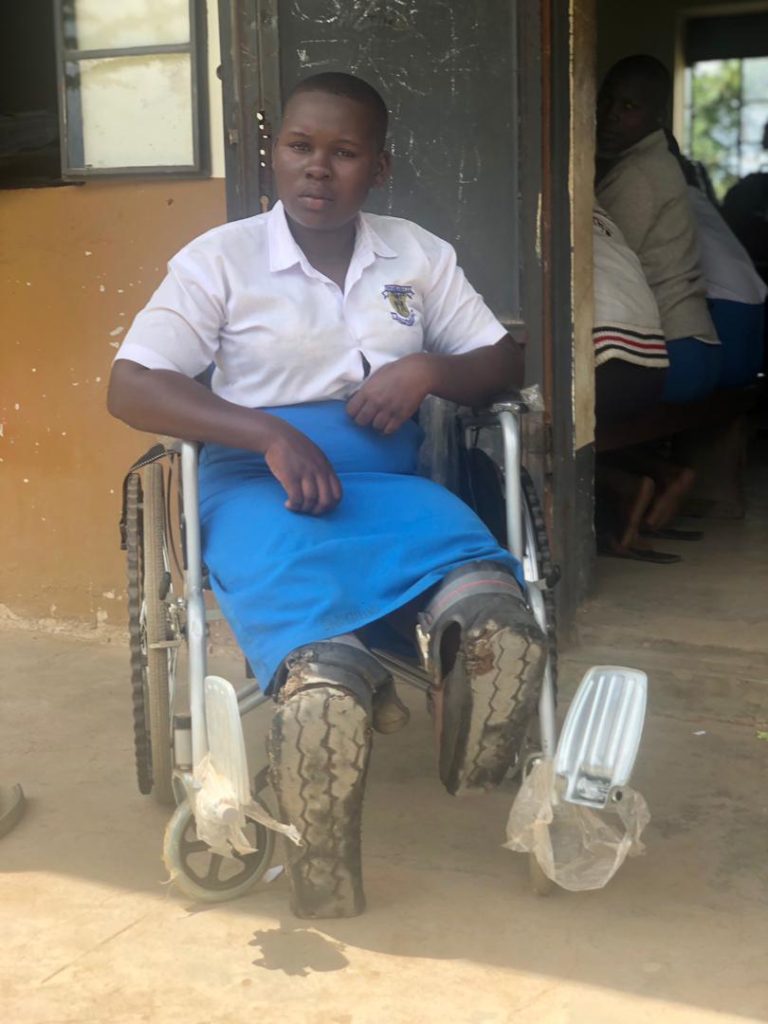 A girl in a wheelchair is sitting outside a classroom. She has kneepads on. 