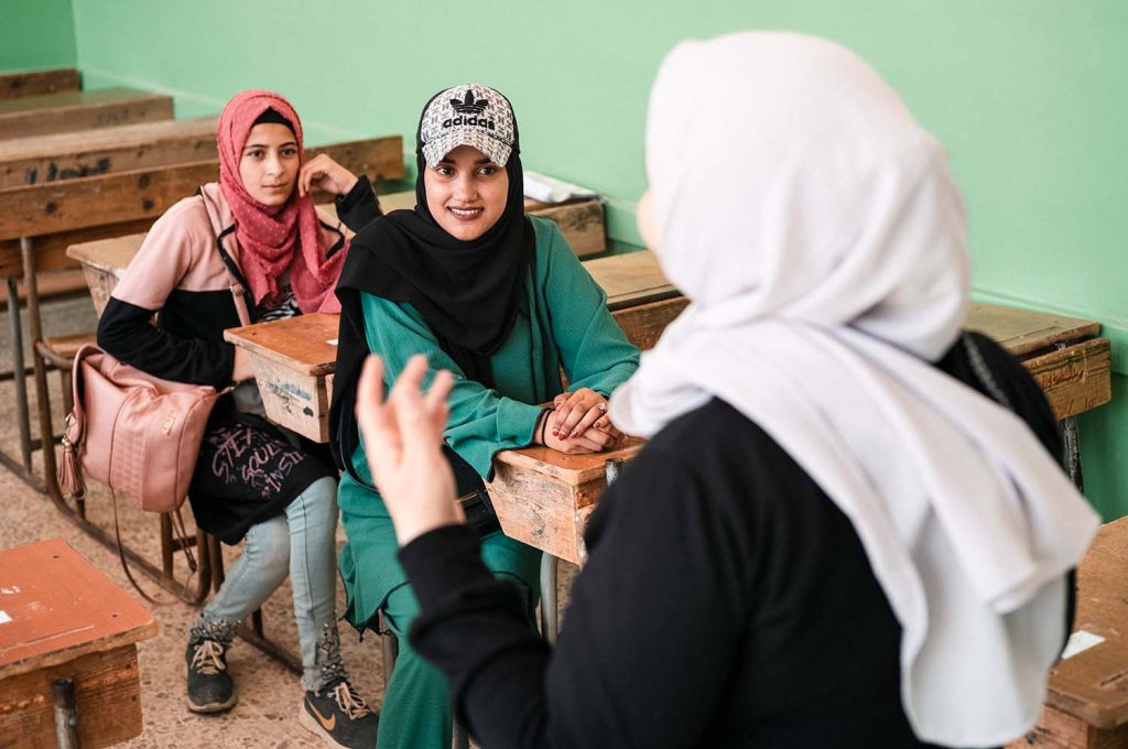 Två tonårsflickor sitter vid sina skrivbord i ett skolklassrum i Syrien och lyssnar på en tredje kvinna med ryggen mot kameran.