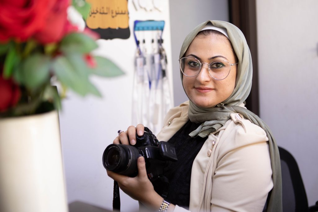 A smiling woman in a hijab holds a camera