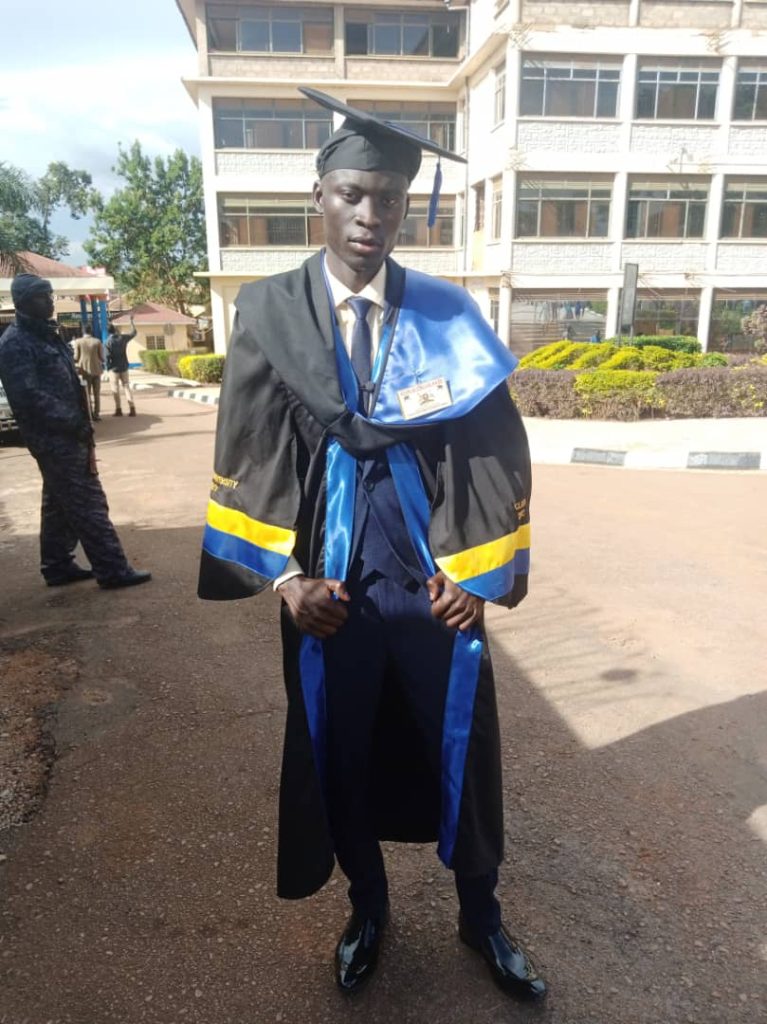 A man in a graduation gown and cap stands outside posing for a photo