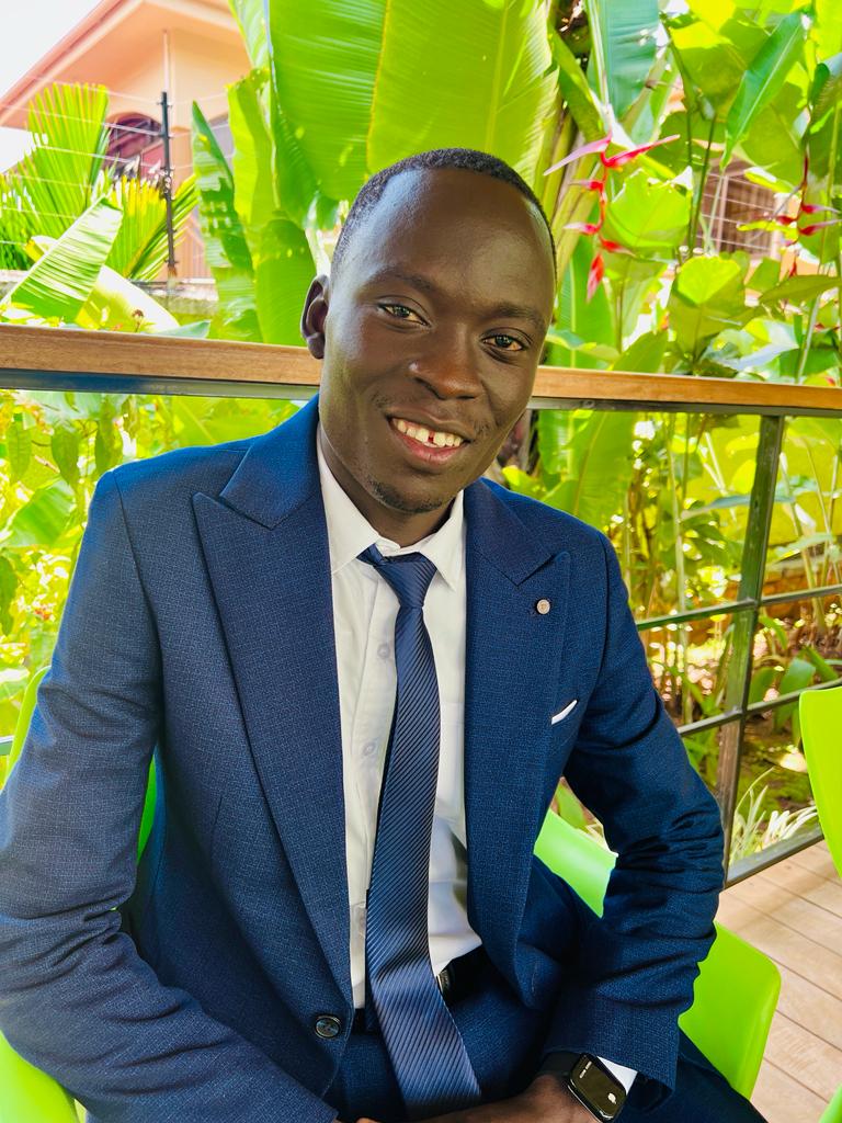 A smiling man in a business suit sits outside in front of foliage.