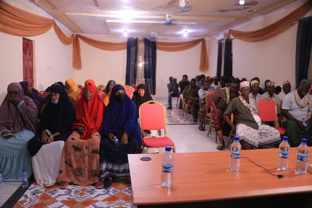 A large number of men and women sit in a room. A table with water bottles is in the foreground